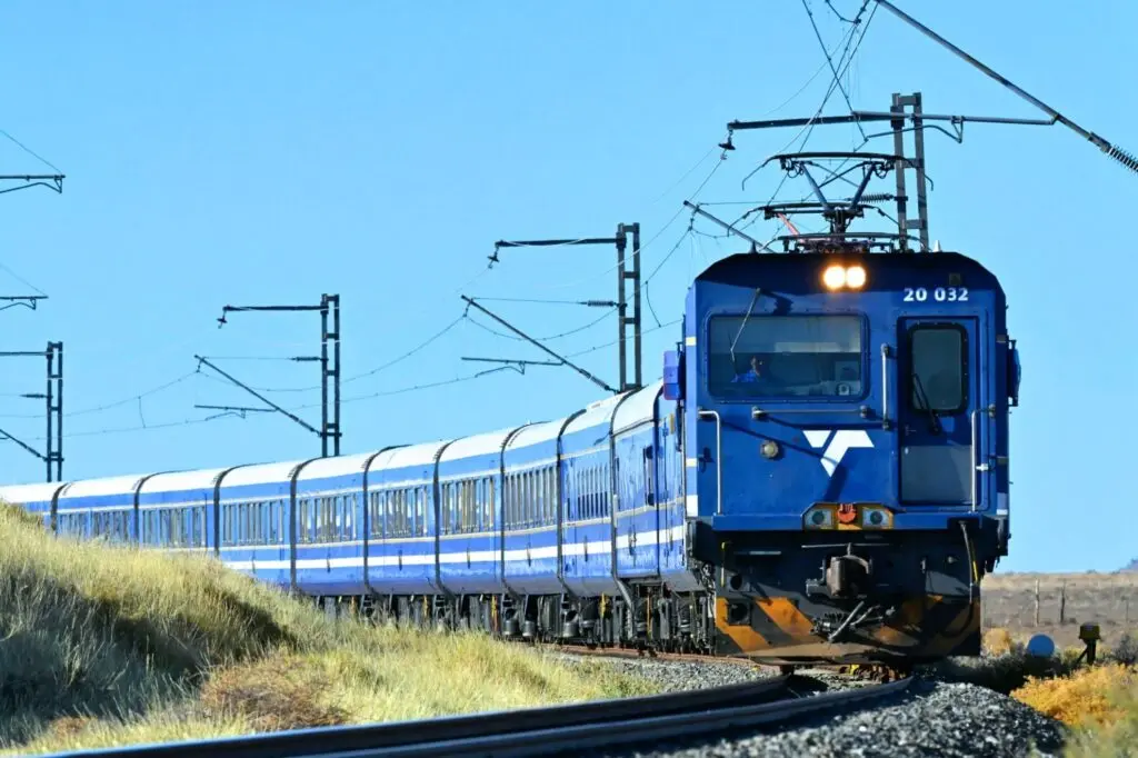 Blue luxury train curving through dry grassland under electric catenary and clear sky